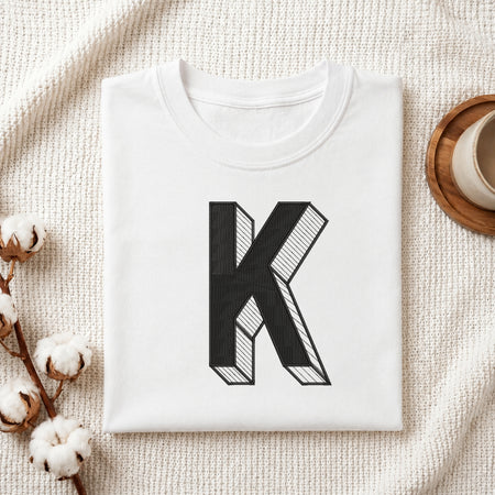 A folded white t-shirt with a large black block letter 'K' printed on the front, styled on a textured cream knit blanket with cotton bolls and a cup in the background.