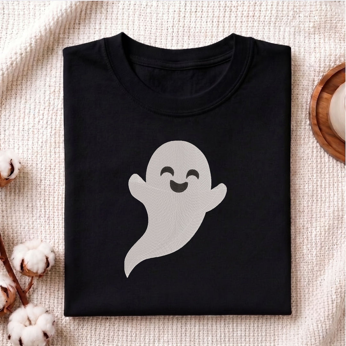 Folded black cotton t-shirt mockup featuring a cute smiling ghost machine embroidery design, styled with cotton stems and a wooden cup.  