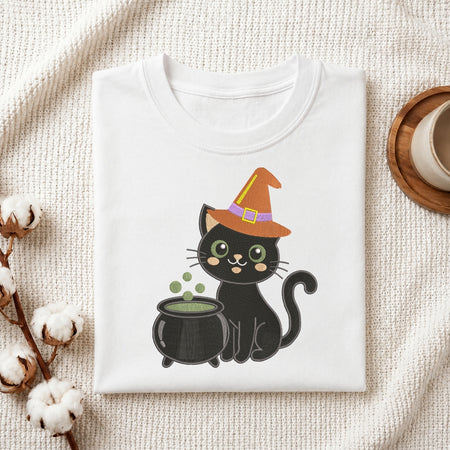 A folded white crewneck t-shirt featuring a cute cartoon graphic of a black cat wearing an orange witch hat next to a cauldron, styled on a textured cream fabric with cotton bolls and a coffee cup.