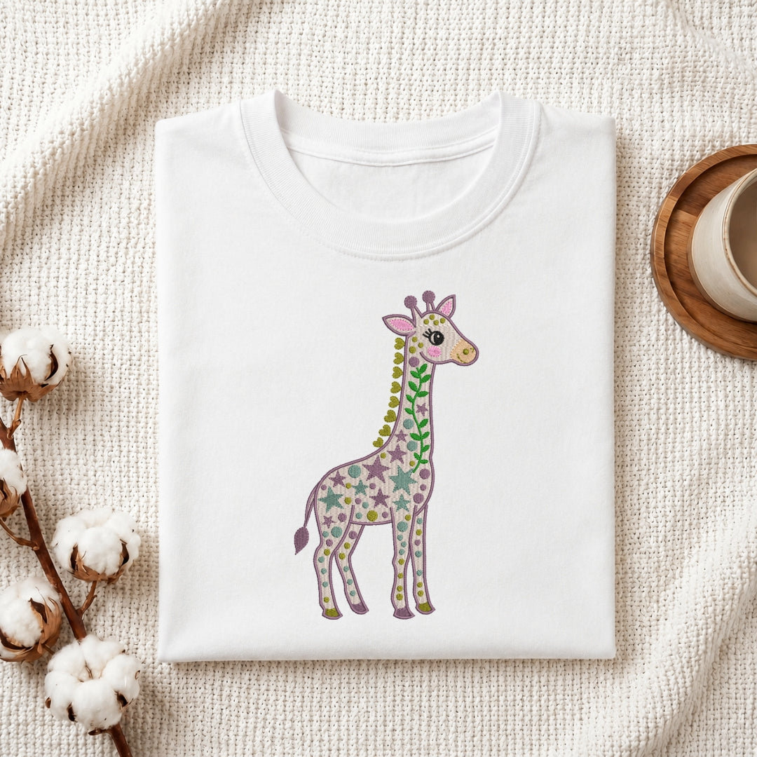 A folded white crewneck t-shirt featuring a colorful giraffe illustration with stars and stripes, styled on a textured cream fabric with cotton bolls and a wooden cup.