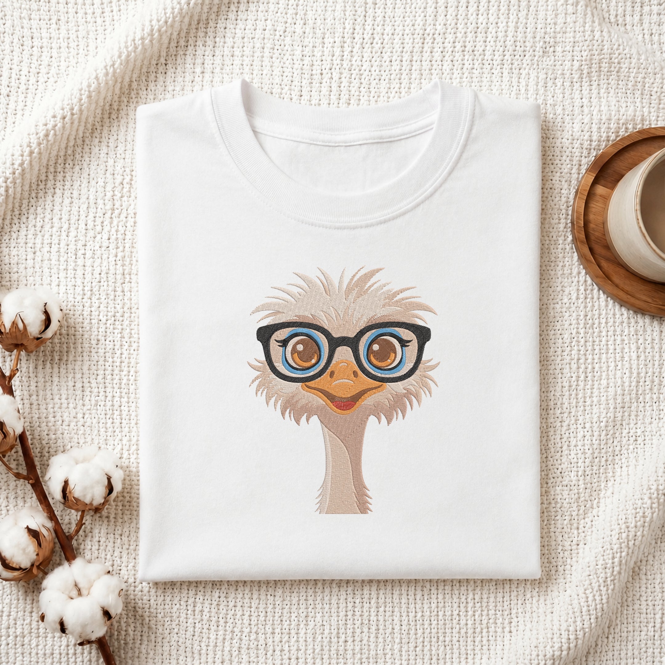 A folded white T-shirt with a cartoon ostrich wearing black-rimmed glasses, displayed on a textured cream fabric with cotton bolls and a small cup as styled props.