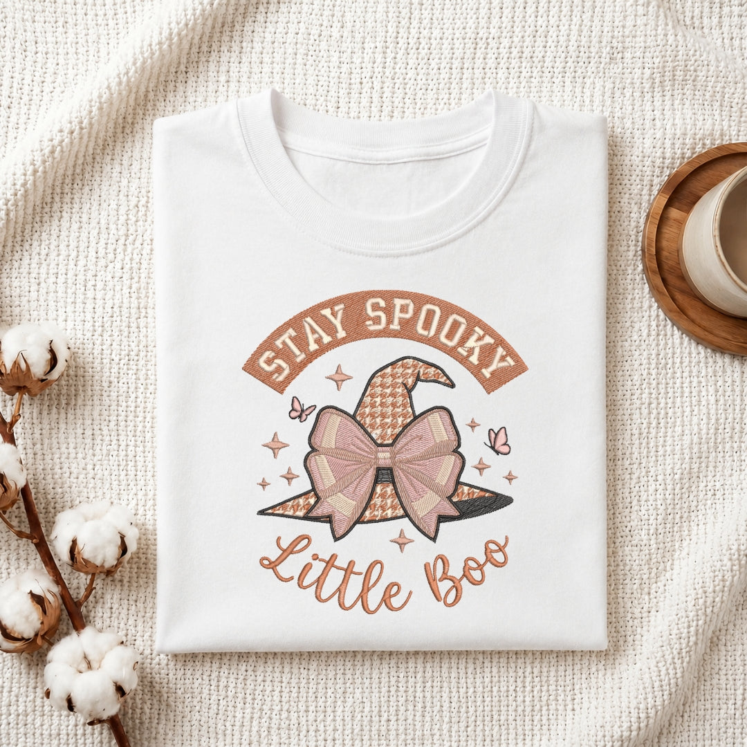 A folded white t-shirt with a 'Stay Spooky Little Boo' graphic featuring a patterned witch hat and bow, styled on a textured cream fabric with cotton bolls and a coffee cup.