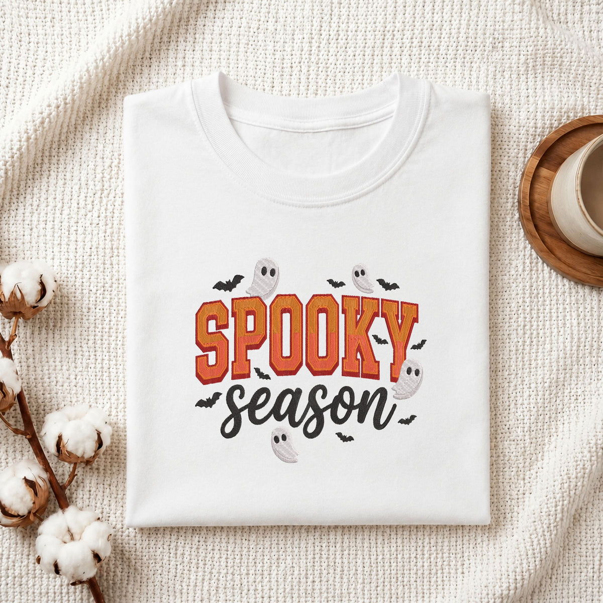 A folded white t-shirt with a 'Spooky Season' graphic print, styled on a textured cream fabric background with cotton bolls and a coffee cup.