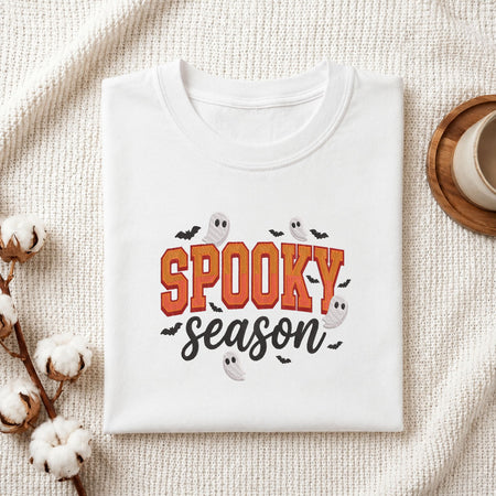 A folded white t-shirt with a 'Spooky Season' graphic print, styled on a textured cream fabric background with cotton bolls and a coffee cup.