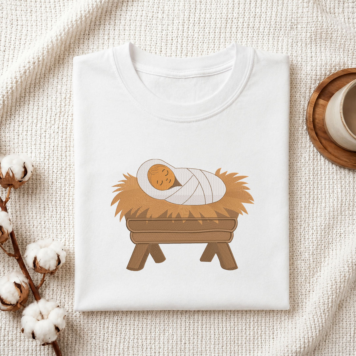 Folded white cotton t-shirt mockup featuring a baby Jesus in a manger machine embroidery design, styled with cotton stems and a wooden cup.