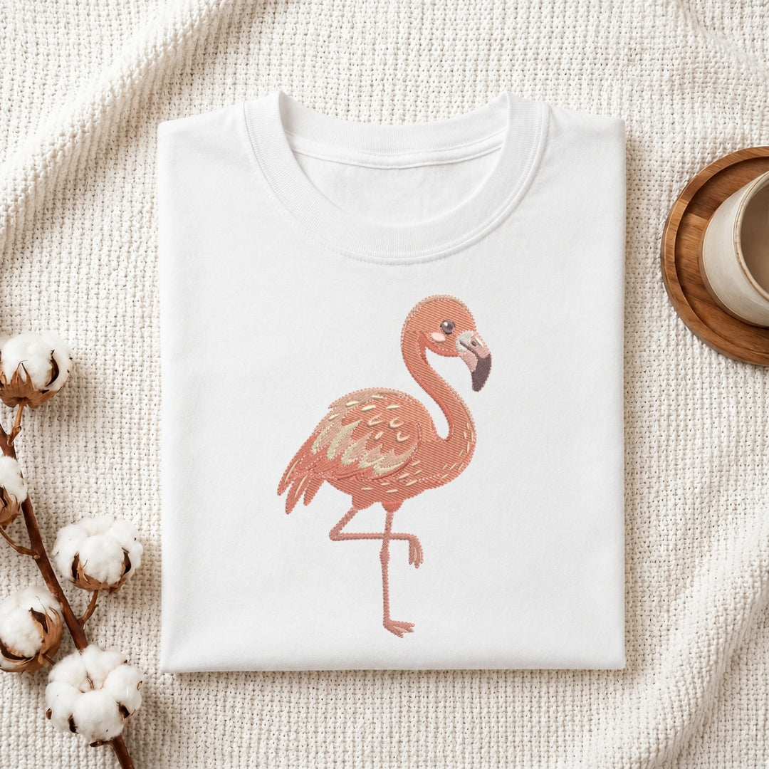 A folded white crewneck t-shirt featuring a printed illustration of a pink flamingo, styled on a textured cream knit blanket with cotton bolls and a wooden cup in the background.