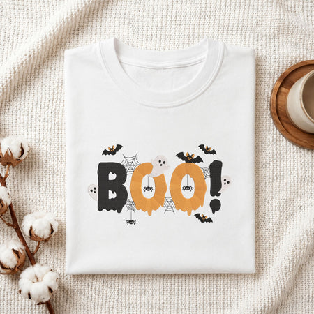 Folded white cotton t-shirt mockup featuring a spooky "BOO!" Halloween machine embroidery design, styled with cotton stems and a wooden cup.