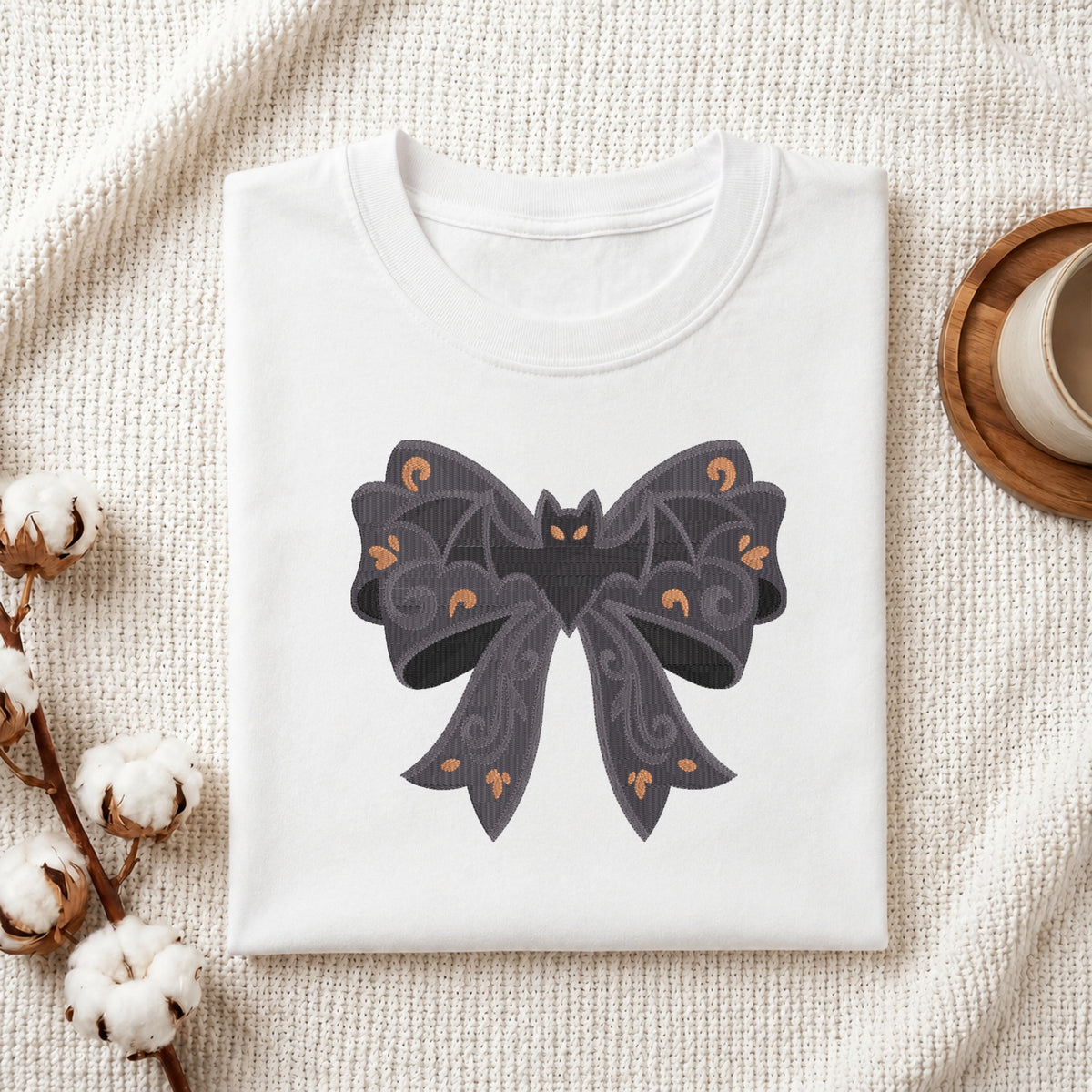 A neatly folded white t-shirt featuring the embroidered gothic bat bow design, displayed on a textured white knit background with cotton stems and a wooden cup.