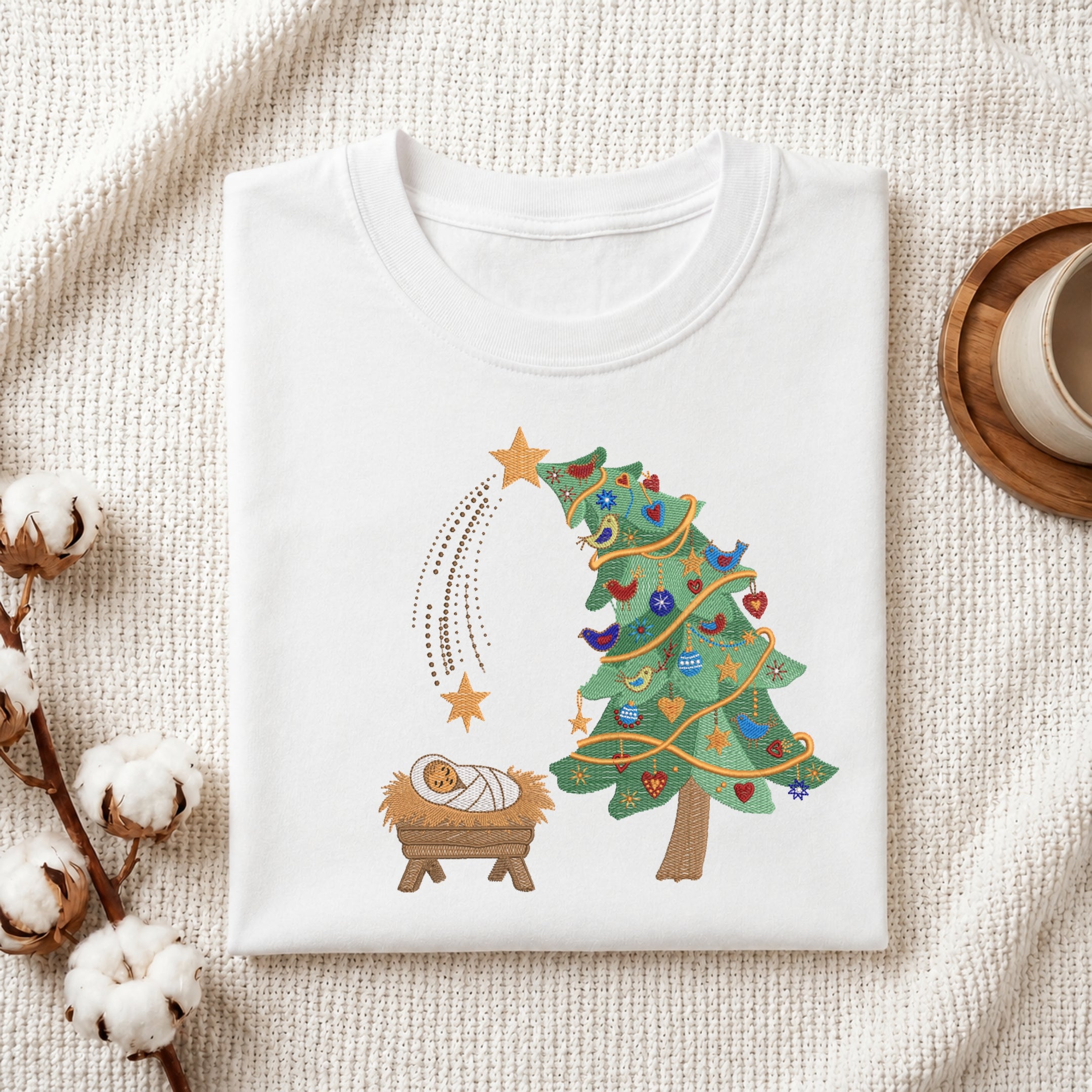 A folded white T-shirt with a Christmas nativity graphic, displayed on a textured cream fabric background with decorative cotton bolls and a cup on a wooden tray.