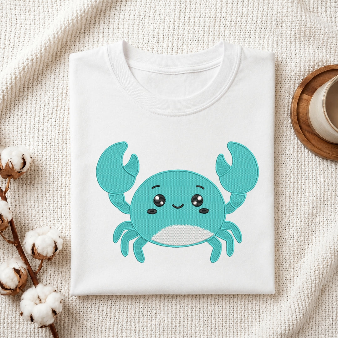A folded white crewneck T-shirt with a cute embroidered-style turquoise crab graphic on the front, styled on a textured cream fabric with cotton bolls and a small cup in the background.