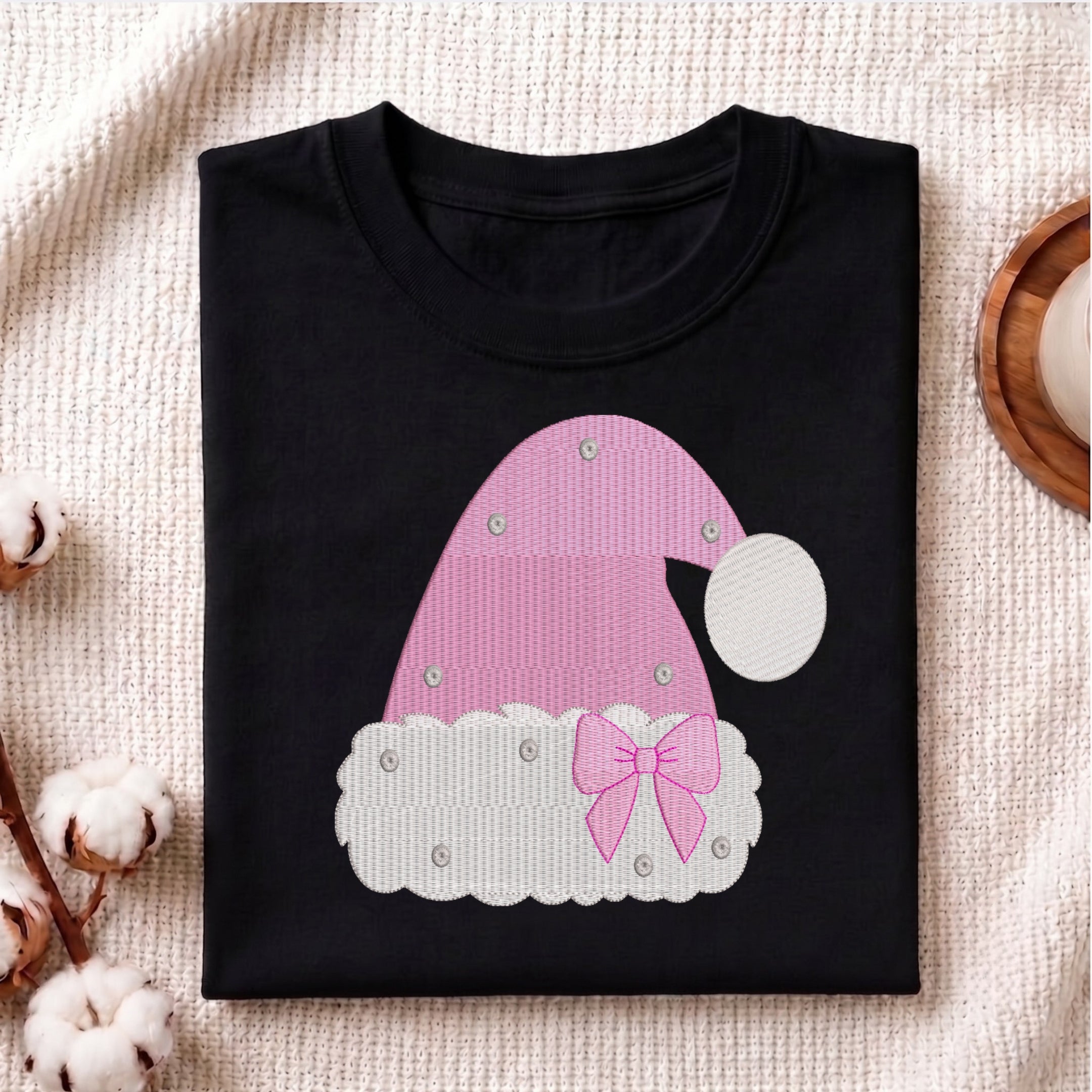 A folded black t-shirt with a pink and white Santa hat applique design, displayed on a textured beige fabric background with decorative cotton bolls.