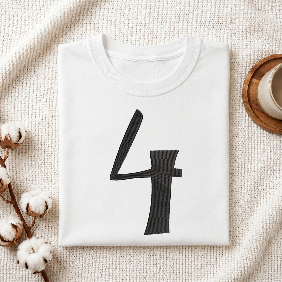 A folded white crewneck t-shirt with a large black number 4 printed on the front, styled on a textured cream fabric with cotton stems and a wooden cup in the background.