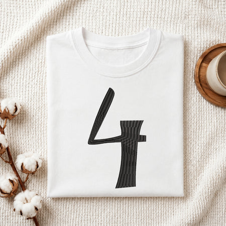 A folded white crewneck t-shirt with a large black number 4 printed on the front, styled on a textured cream fabric with cotton stems and a wooden cup in the background.