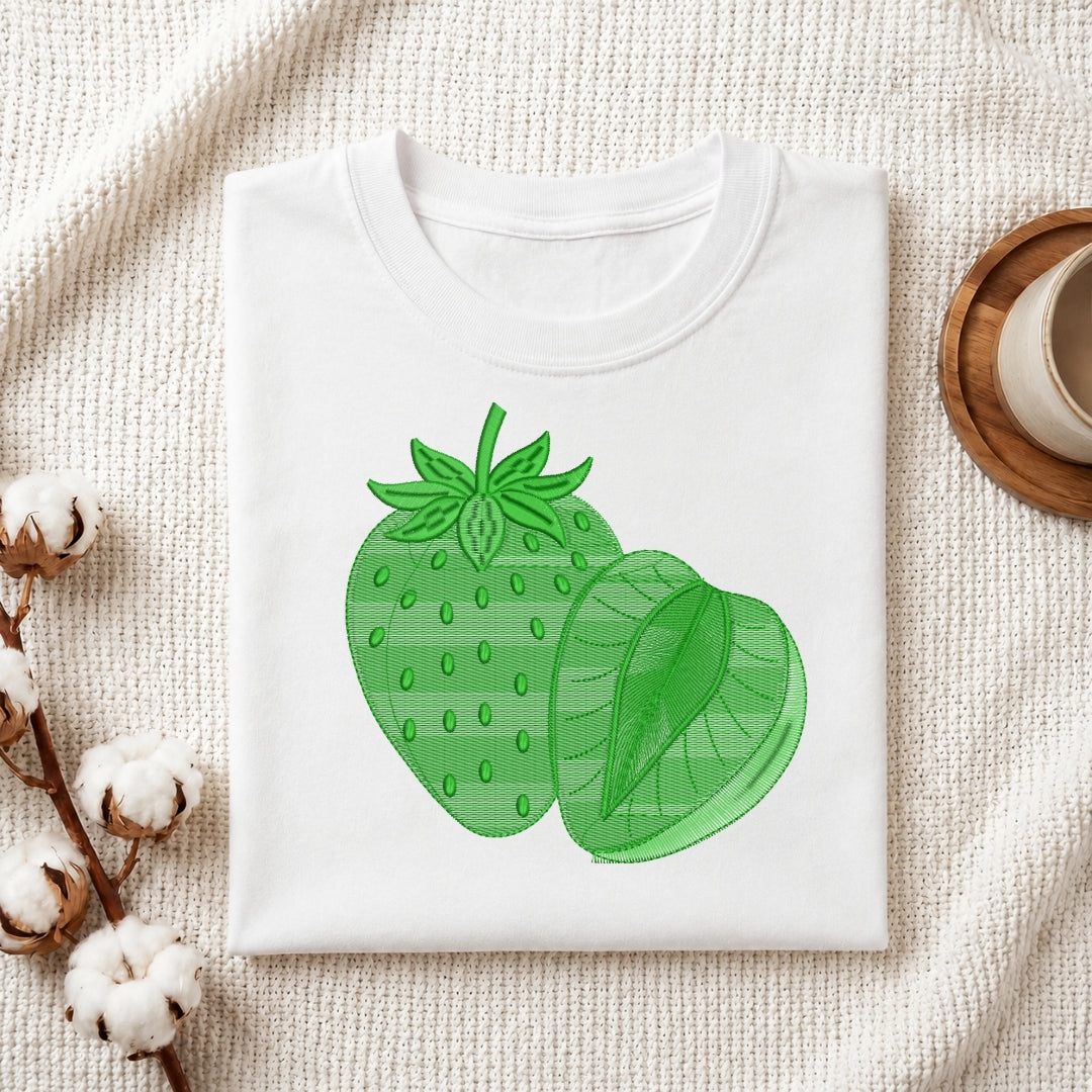 A folded white crewneck t-shirt featuring a green strawberry and strawberry slice graphic on the front, styled on a textured cream fabric with cotton bolls and a wooden cup in the background.