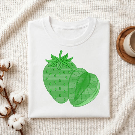 A folded white crewneck t-shirt featuring a green strawberry and strawberry slice graphic on the front, styled on a textured cream fabric with cotton bolls and a wooden cup in the background.
