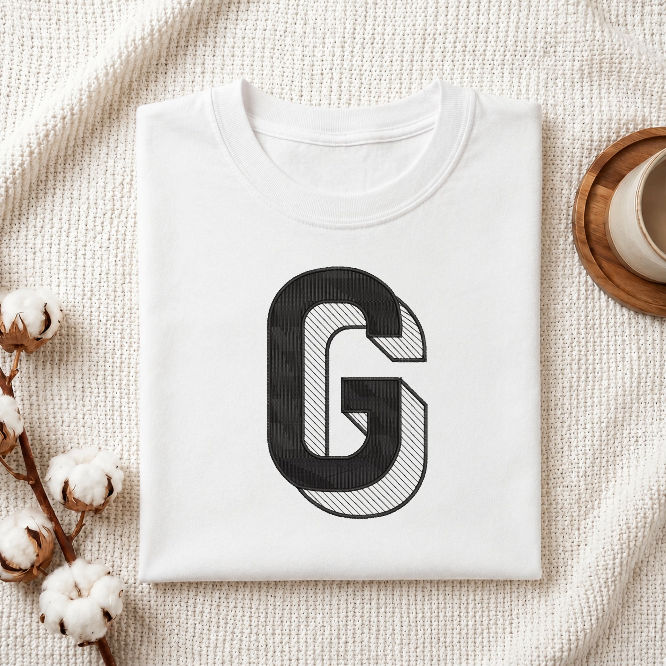 A folded white crewneck t-shirt with a large black and white striped letter 'G' graphic on the front, styled on a textured cream knit blanket with cotton bolls and a wooden cup in the background.