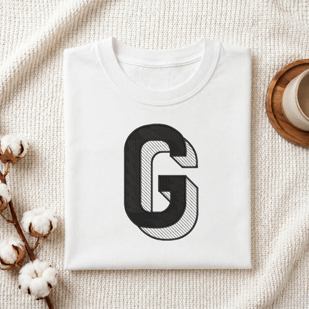A folded white crewneck t-shirt with a large black and white striped letter 'G' graphic on the front, styled on a textured cream knit blanket with cotton bolls and a wooden cup in the background.