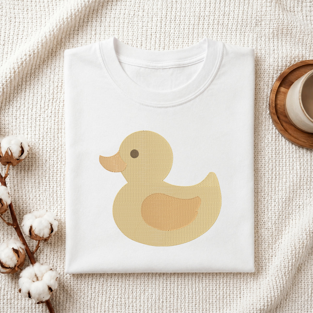 A folded white T-shirt with a large yellow duck graphic on the front, styled on a textured cream fabric with cotton bolls and a wooden cup edge visible.