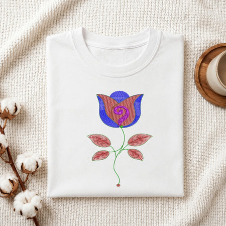 A folded white crewneck t-shirt with a colorful embroidered rose design, displayed on a textured cream fabric background with decorative cotton bolls and a wooden cup.