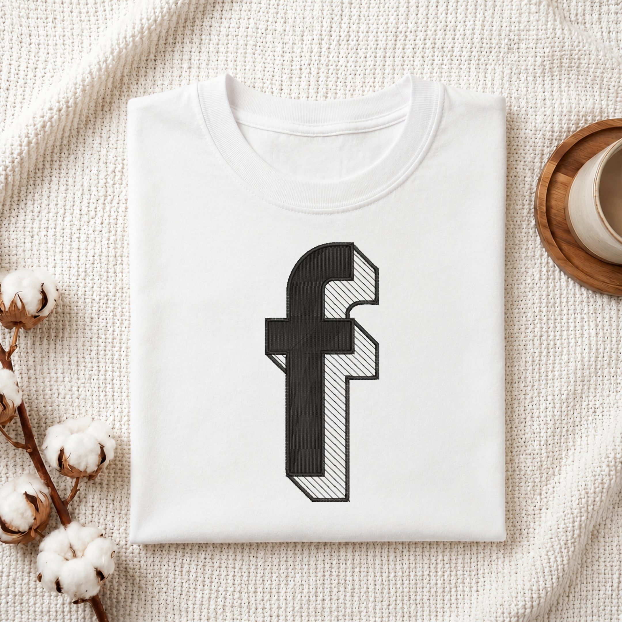 A neatly folded white crewneck t-shirt featuring a stitched 3D block lowercase letter "f" design, styled with cotton branches and a wooden coaster.