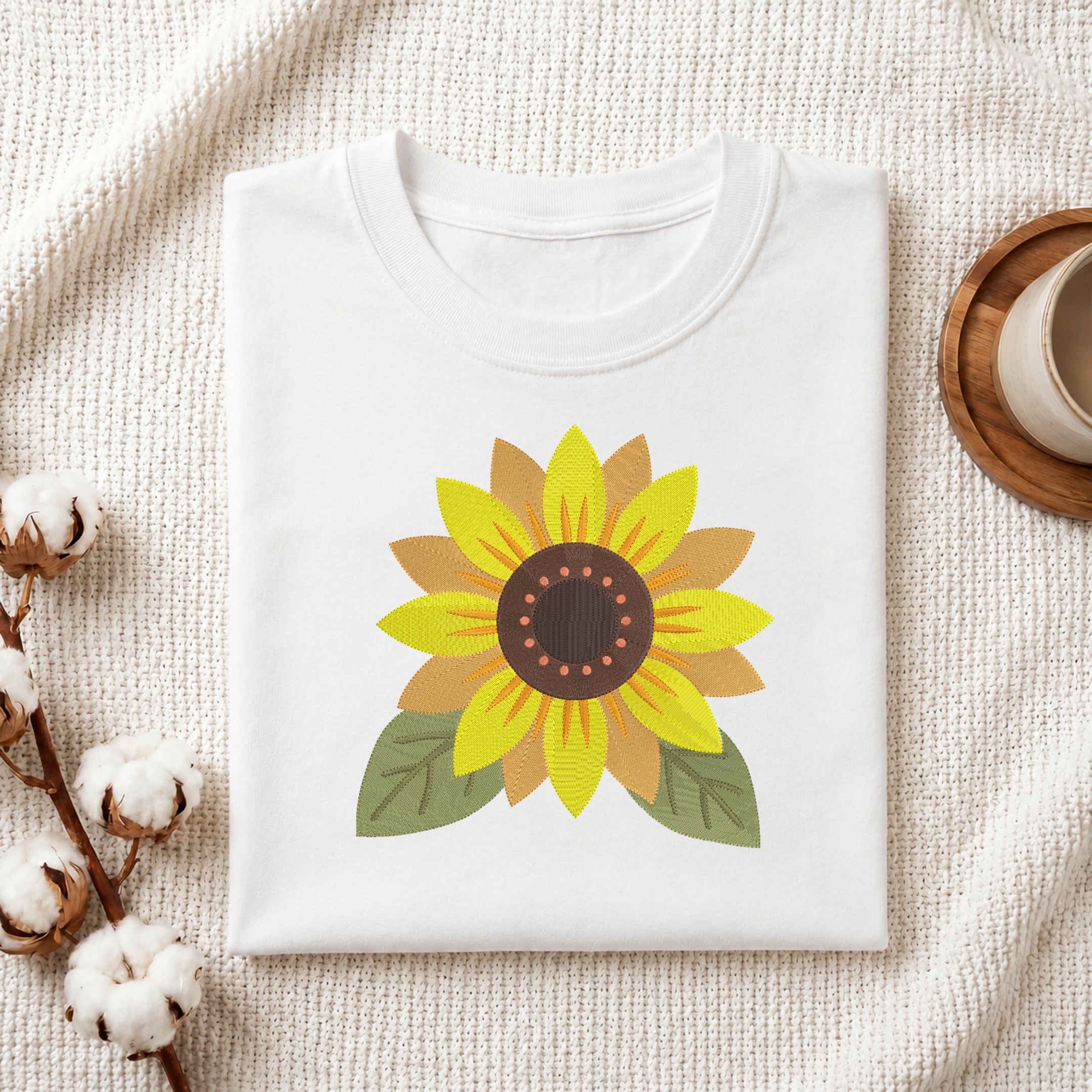 A folded white crewneck t-shirt featuring a colorful sunflower graphic on the front, styled on a textured cream fabric background with cotton bolls and a wooden cup.