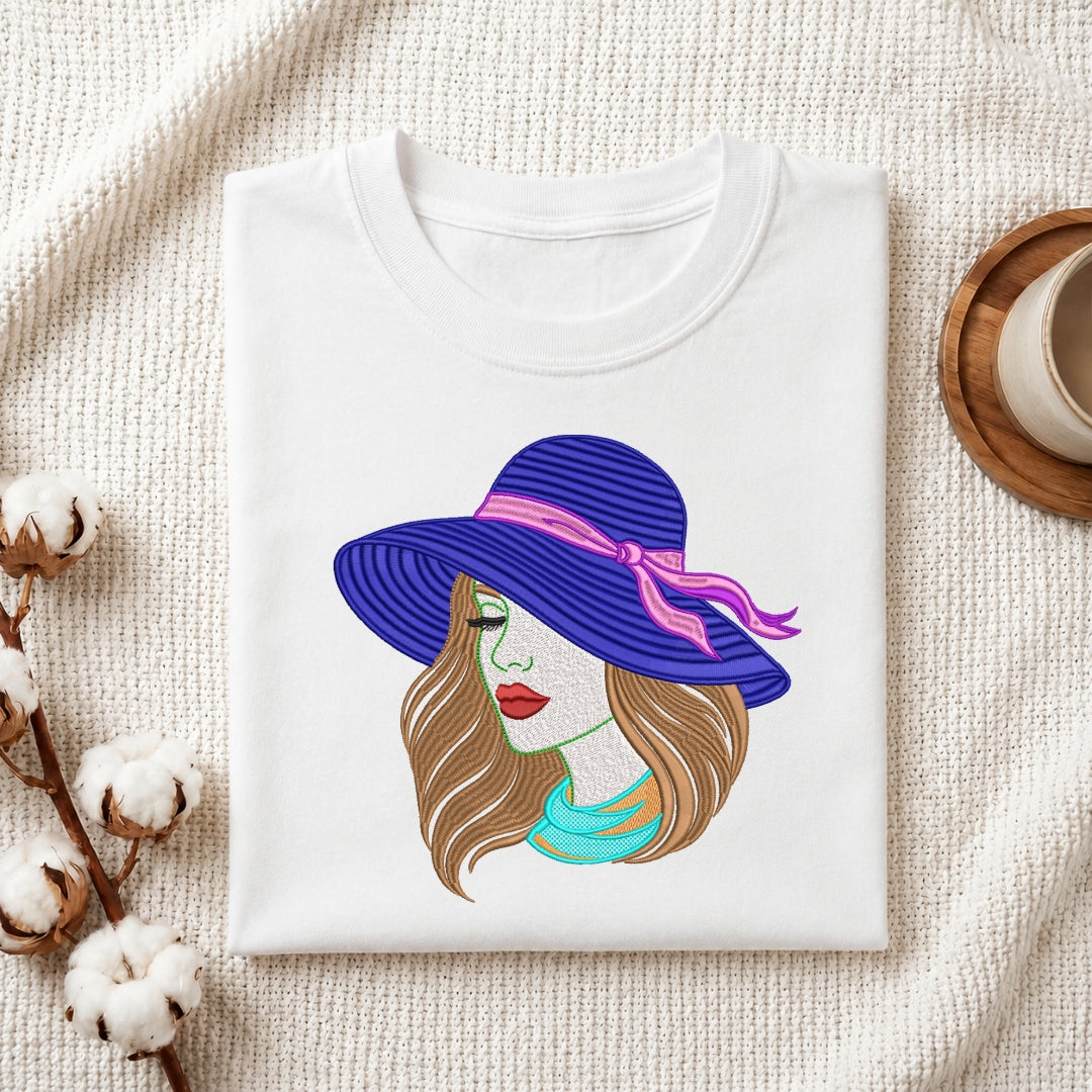 A folded white crewneck t-shirt displayed on a textured cream fabric background, decorated with cotton bolls and a wooden cup. The t-shirt features a colorful graphic illustration of a woman wearing a purple striped sunhat with a pink ribbon and a turquoise top.