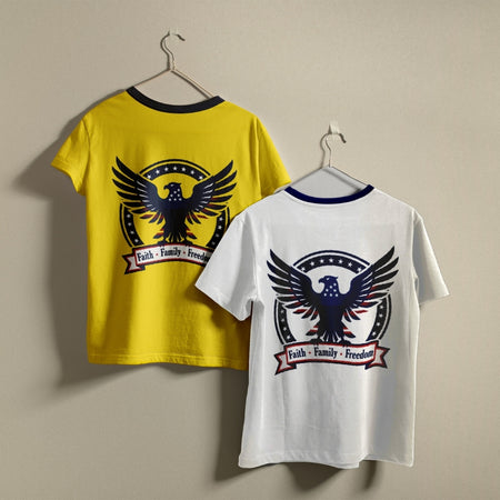 Two t-shirts hanging on hangers, one yellow and one white, both printed with a patriotic eagle graphic and the text "Faith • Family • Freedom".