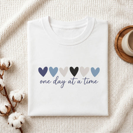 A folded white t-shirt with a graphic of six colored hearts and the text 'one day at a time' in navy script, displayed on a textured cream fabric background with cotton bolls and a cup in the corners.