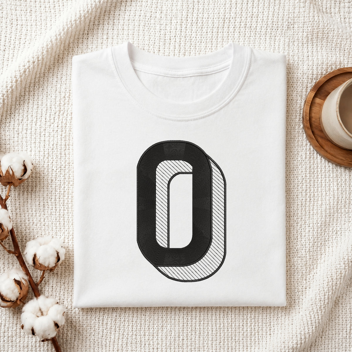 A folded white crewneck t-shirt with a large black graphic letter 'O' on the front, styled on a textured cream blanket with cotton bolls and a wooden cup in the background.