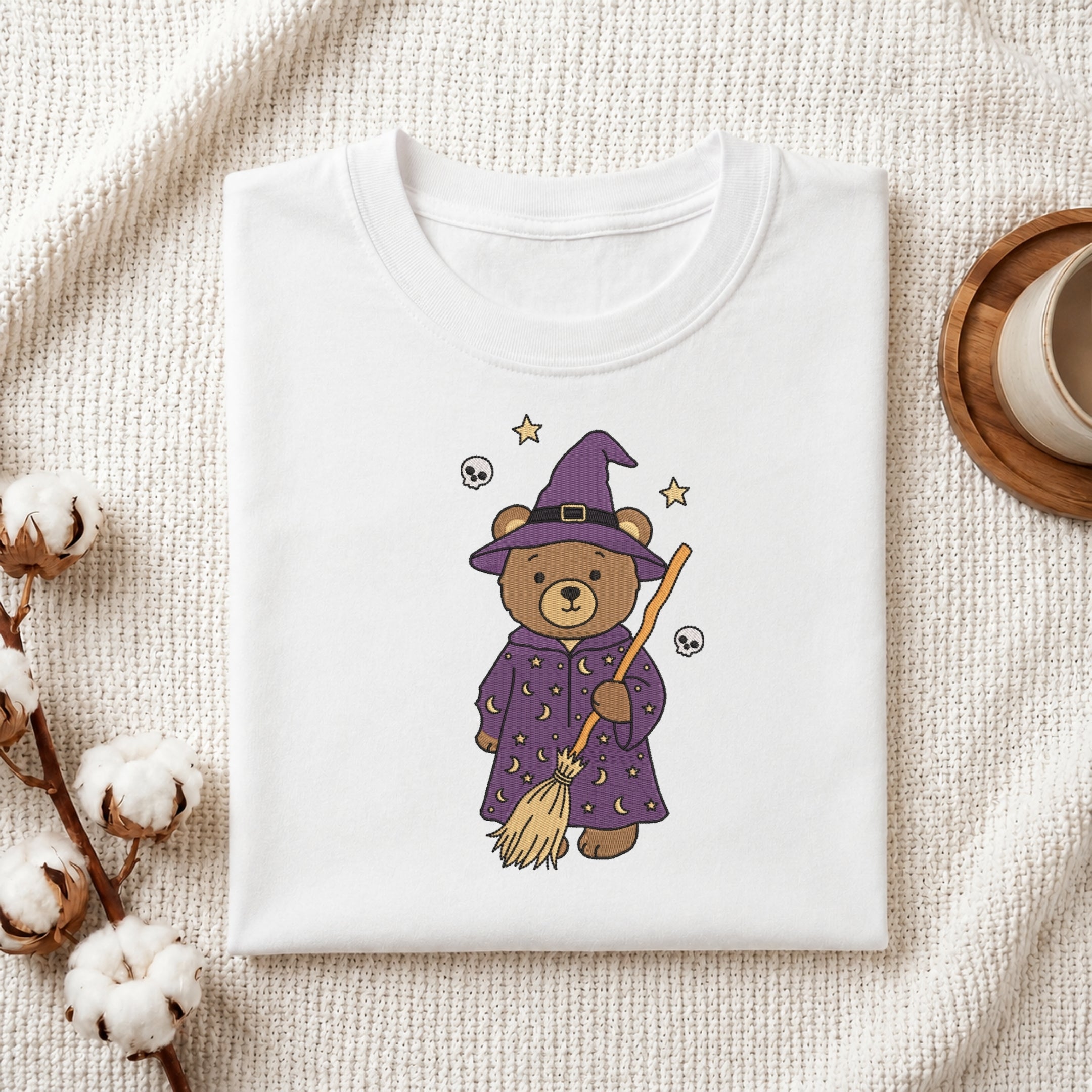 A folded white T-shirt displayed on a textured cream fabric background, featuring a cute cartoon bear dressed as a witch in a purple robe and hat, holding a broomstick with small star and skull details.