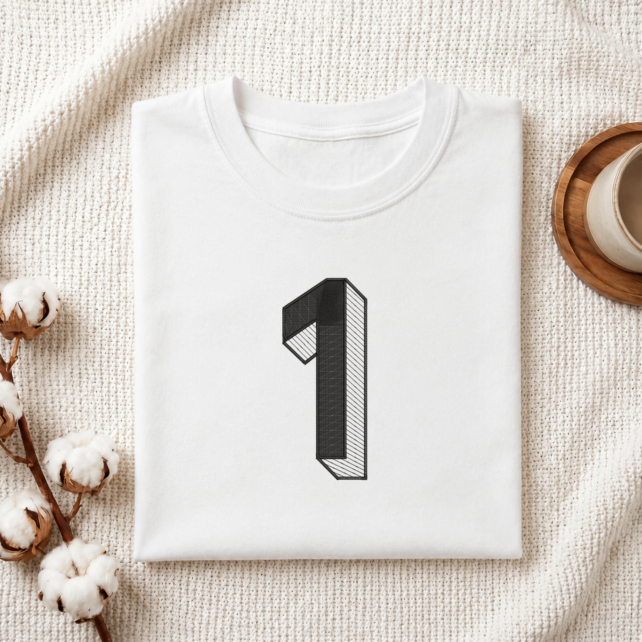 A folded white t-shirt with a black 3D-style number '1' graphic on the chest, styled in a flat lay on a textured cream fabric with cotton bolls and a wooden cup.