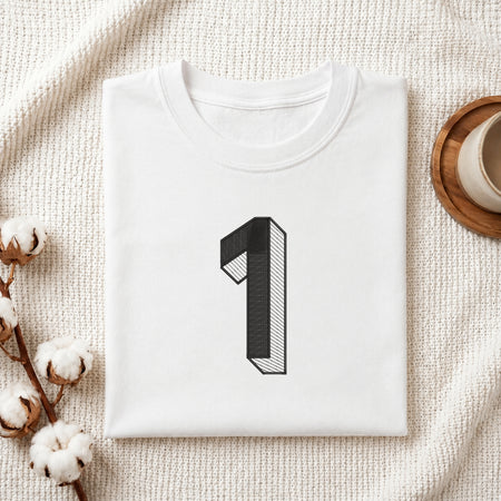 A folded white t-shirt with a black 3D-style number '1' graphic on the chest, styled in a flat lay on a textured cream fabric with cotton bolls and a wooden cup.