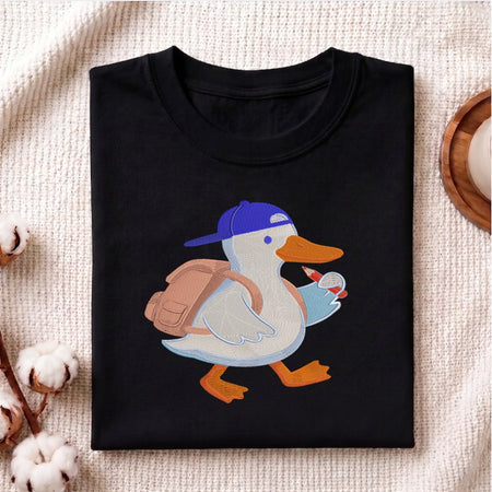 A folded black t-shirt with a cartoon duck graphic wearing a blue cap and brown backpack, displayed on a textured beige fabric background with cotton bolls and a wooden coaster.