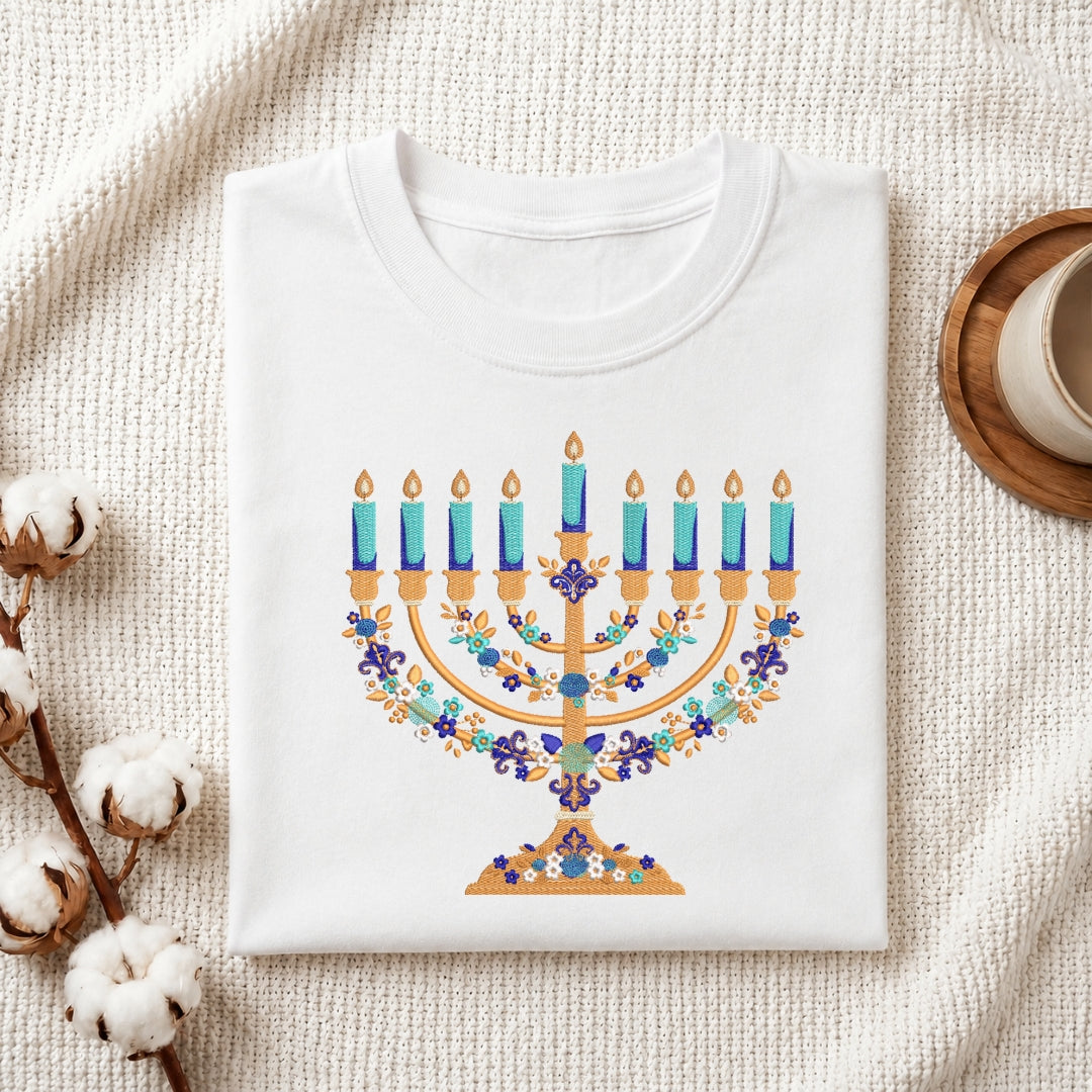 A folded white crewneck t-shirt featuring a colorful printed Hanukkah menorah design, styled on a textured cream fabric background with cotton bolls and a wooden cup.