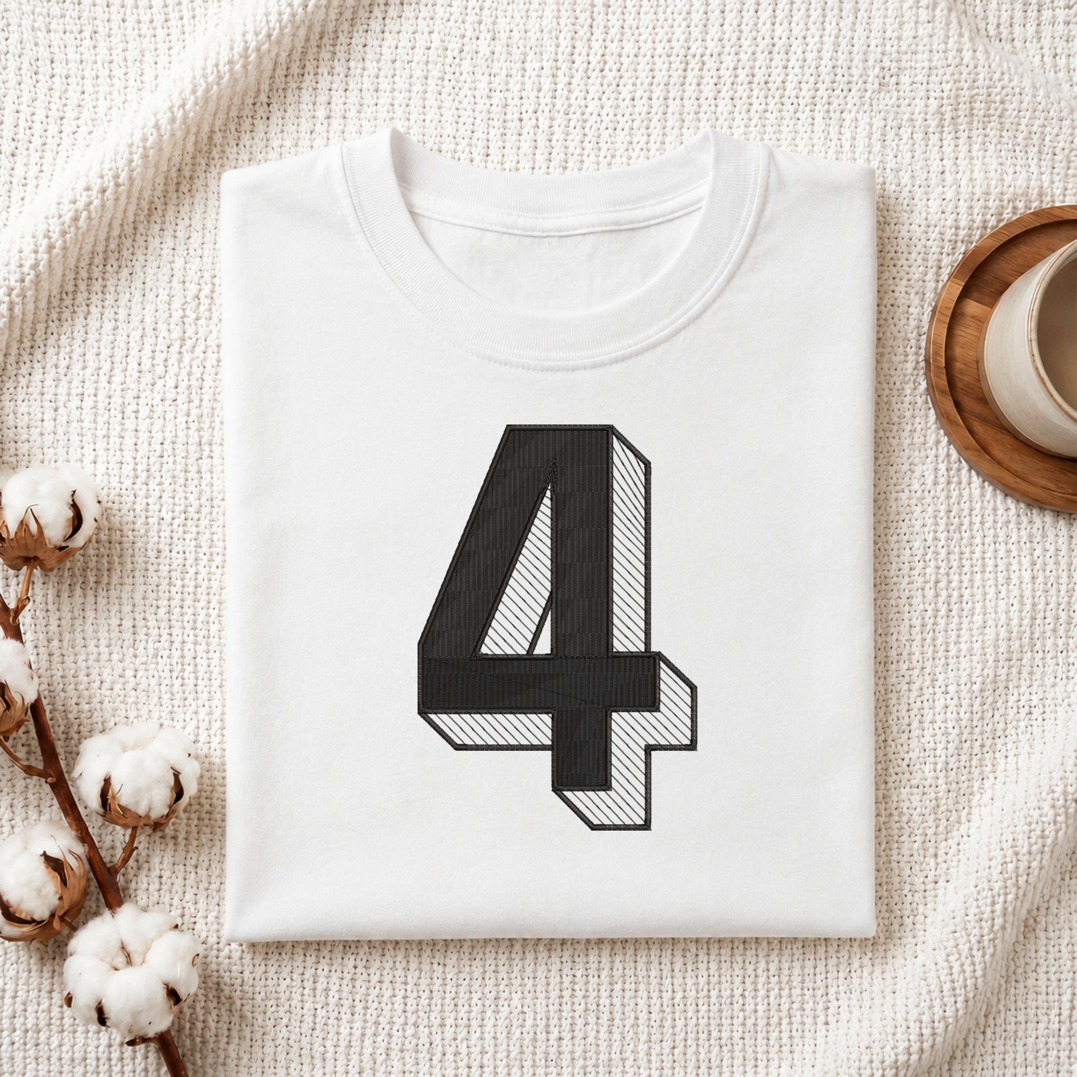 A neatly folded white crewneck t-shirt featuring a stitched 3D block number "4" design, styled with cotton branches and a wooden coaster.
