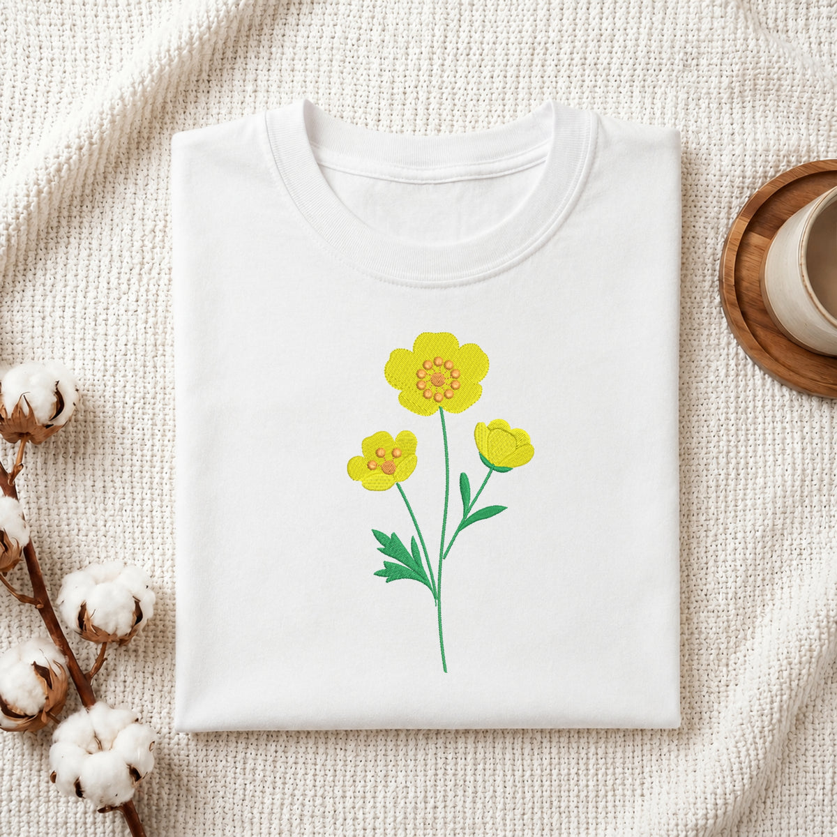 A folded white crewneck t-shirt with a yellow flower graphic, styled on a textured cream fabric with cotton bolls and a wooden cup in the background.