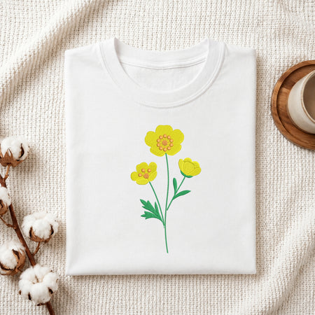 A folded white crewneck t-shirt with a yellow flower graphic, styled on a textured cream fabric with cotton bolls and a wooden cup in the background.