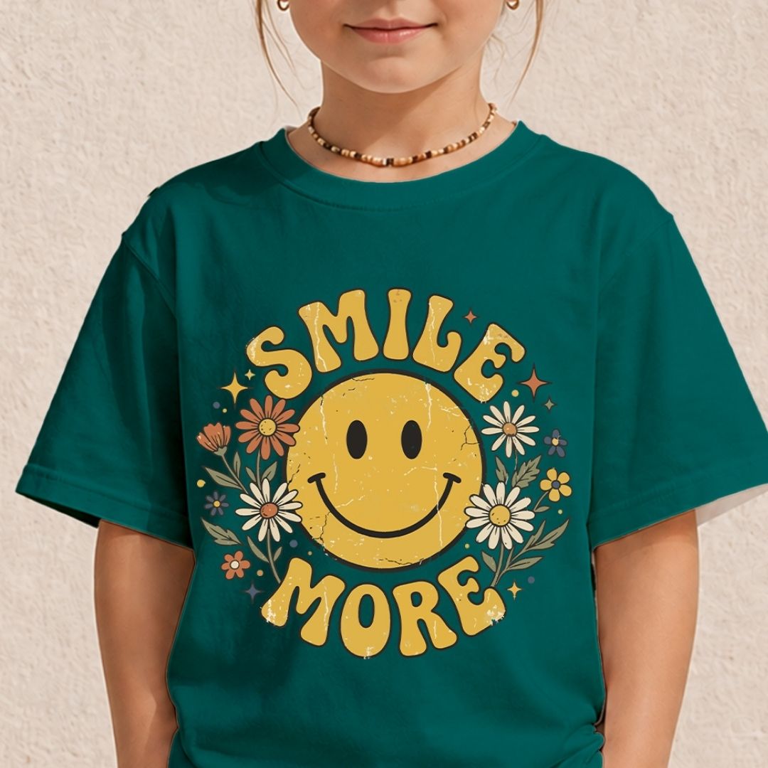A young child wearing a dark teal t-shirt printed with a cute vintage yellow smiley face graphic with colorful daisies and motivational text.