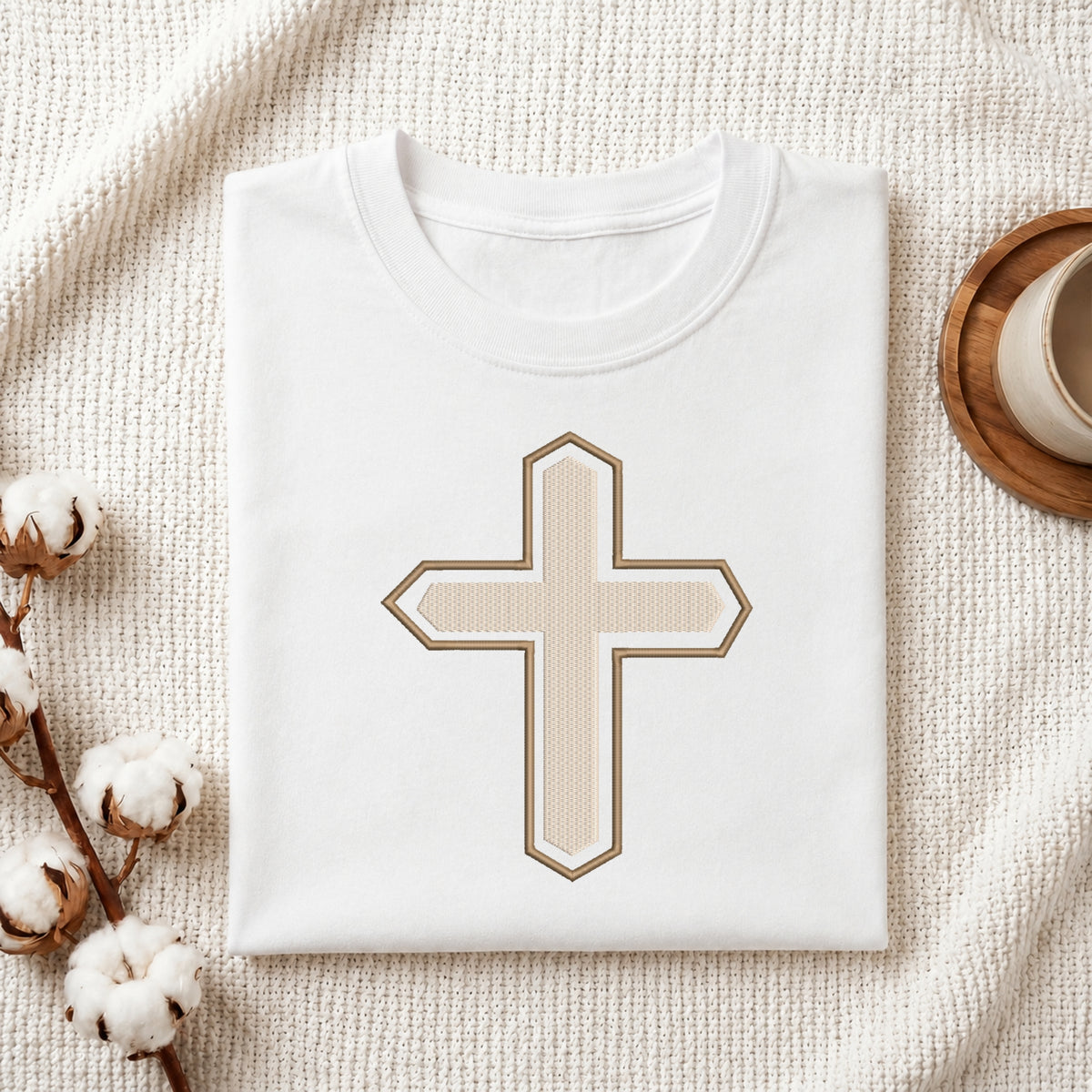 A folded white crewneck t-shirt with a central beige cross graphic outlined in brown, styled on a textured cream blanket with cotton bolls and a cup in the background.