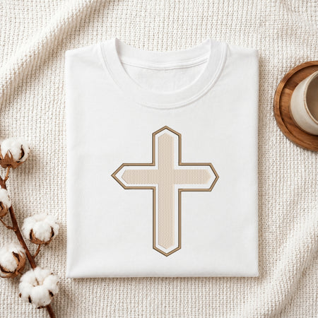 A folded white crewneck t-shirt with a central beige cross graphic outlined in brown, styled on a textured cream blanket with cotton bolls and a cup in the background.