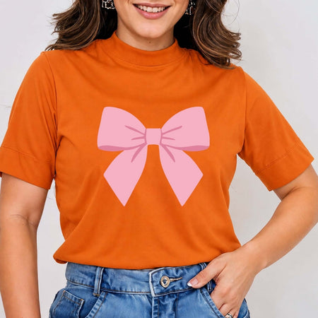 A woman wearing a bright orange short-sleeve mock neck t-shirt with a large pink bow graphic printed on the front, paired with blue jeans.
