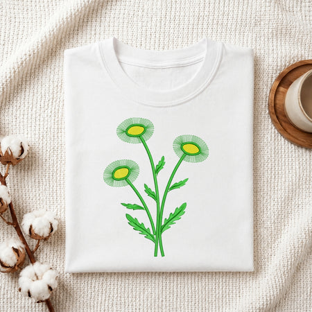 A folded white crewneck t-shirt featuring a green and yellow daisy flower graphic on the front, styled on a textured cream fabric with cotton bolls and a wooden cup in the background.