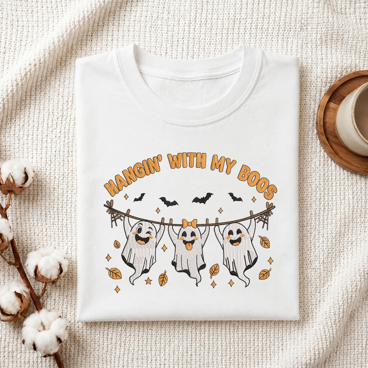 A folded white t-shirt with a Halloween graphic print featuring three ghosts on a clothesline with the text 'HANGIN' WITH MY BOOS', styled on a textured cream fabric background with cotton bolls and a cup.