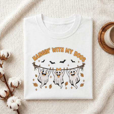 A folded white t-shirt with a Halloween graphic print featuring three ghosts on a clothesline with the text 'HANGIN' WITH MY BOOS', styled on a textured cream fabric background with cotton bolls and a cup.