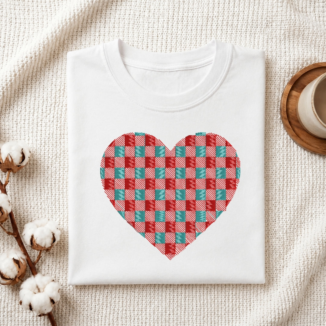 A folded white crewneck t-shirt with a red and teal checkered heart graphic on the front, styled on a textured cream fabric with cotton bolls and a wooden cup.