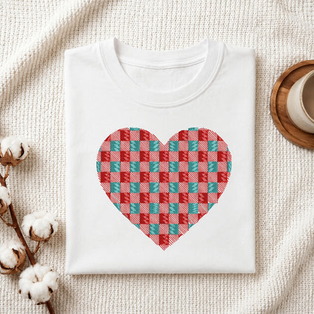 A folded white crewneck t-shirt with a red and teal checkered heart graphic on the front, styled on a textured cream fabric with cotton bolls and a wooden cup.