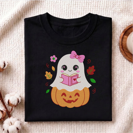 A folded black t-shirt with a cute cartoon graphic of a white ghost reading a pink book on top of an orange jack-o'-lantern, styled on a textured beige fabric with cotton bolls.