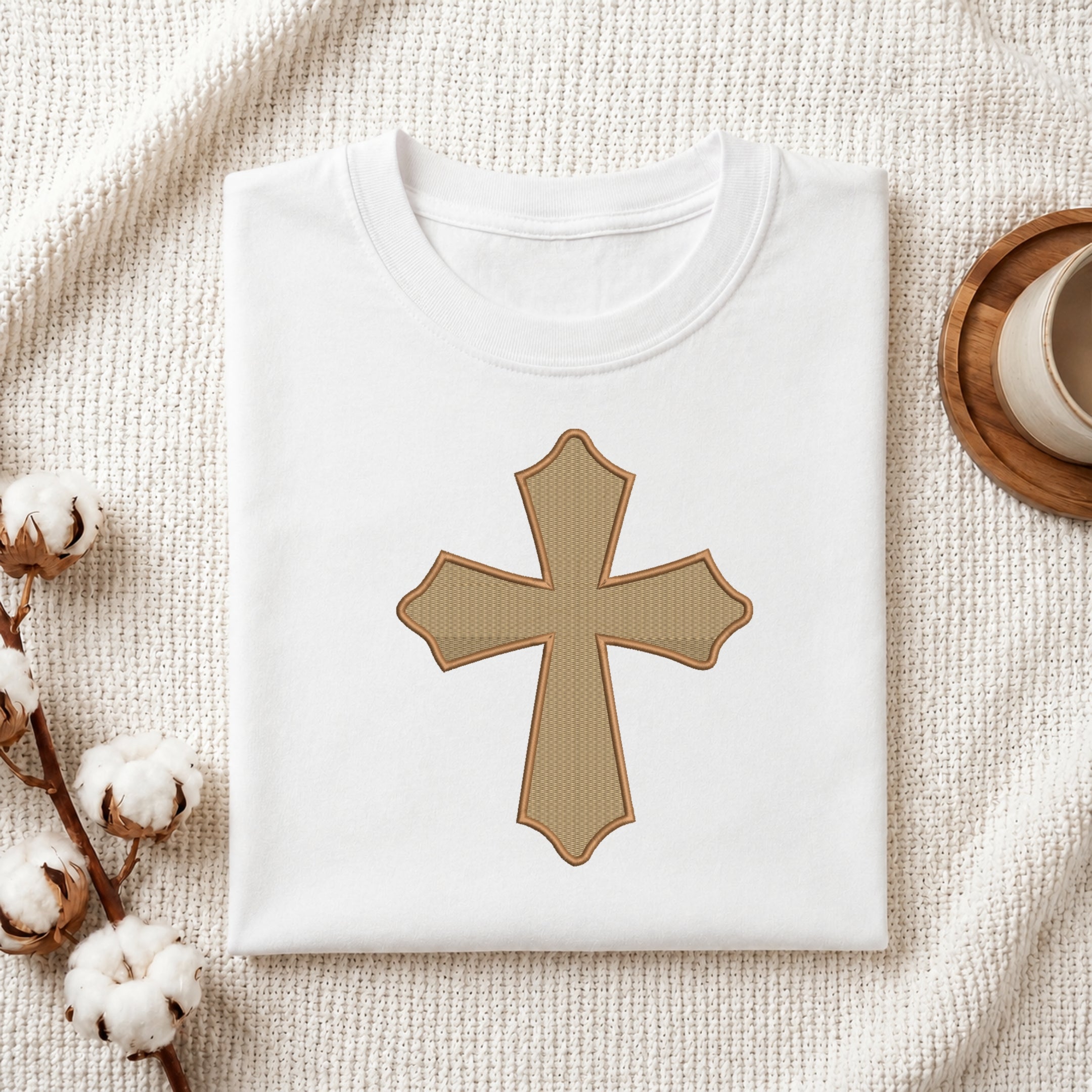 A folded white crewneck t-shirt with a tan cross graphic on the front, styled on a textured cream blanket with cotton bolls and a wooden cup in the background.