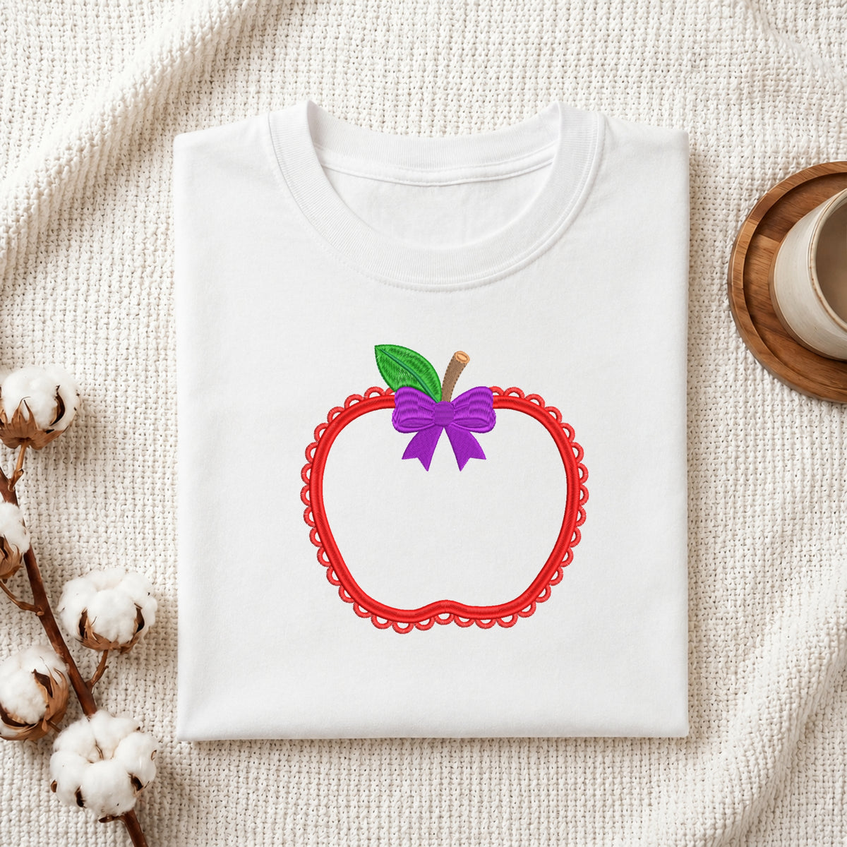 A folded white t-shirt with a graphic of a red scalloped apple outline, a green leaf, and a purple bow, displayed on a textured cream fabric with cotton bolls and a cup in the background.