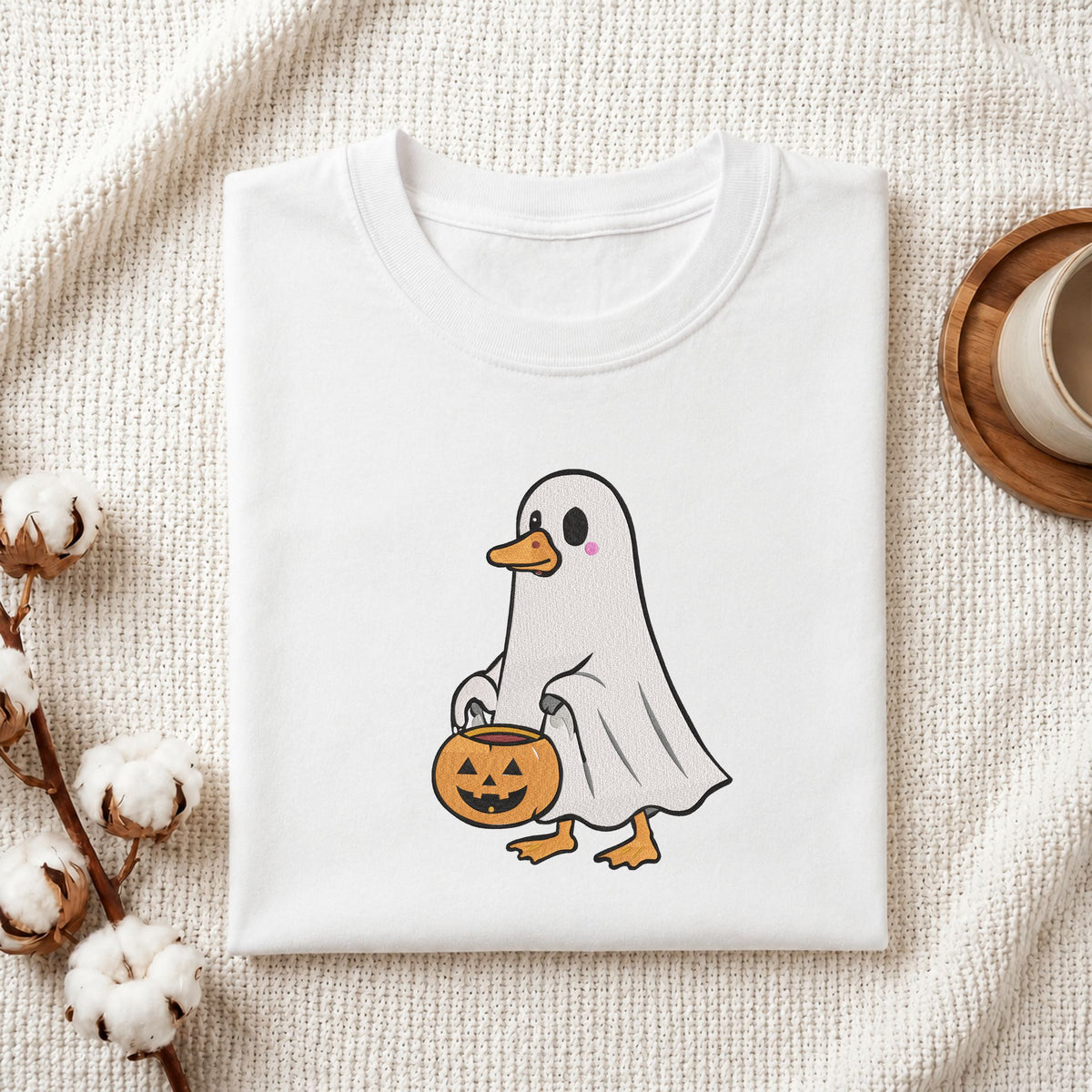 Folded white cotton t-shirt mockup featuring a cute duck in a ghost costume holding a trick-or-treat bucket machine embroidery design, styled with cotton stems and a wooden cup